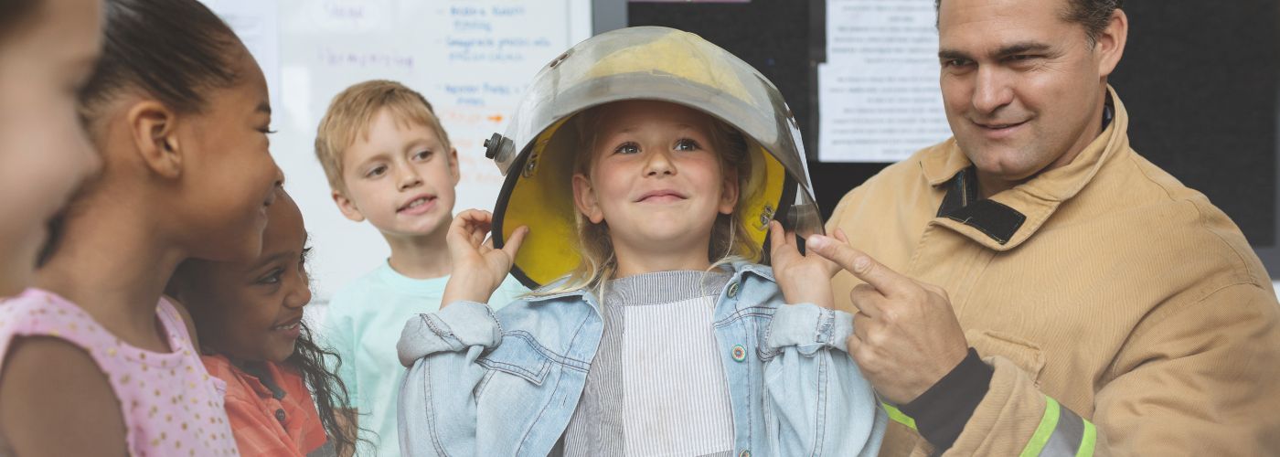 child wearing fireman helmet surrounded by children and a fireman